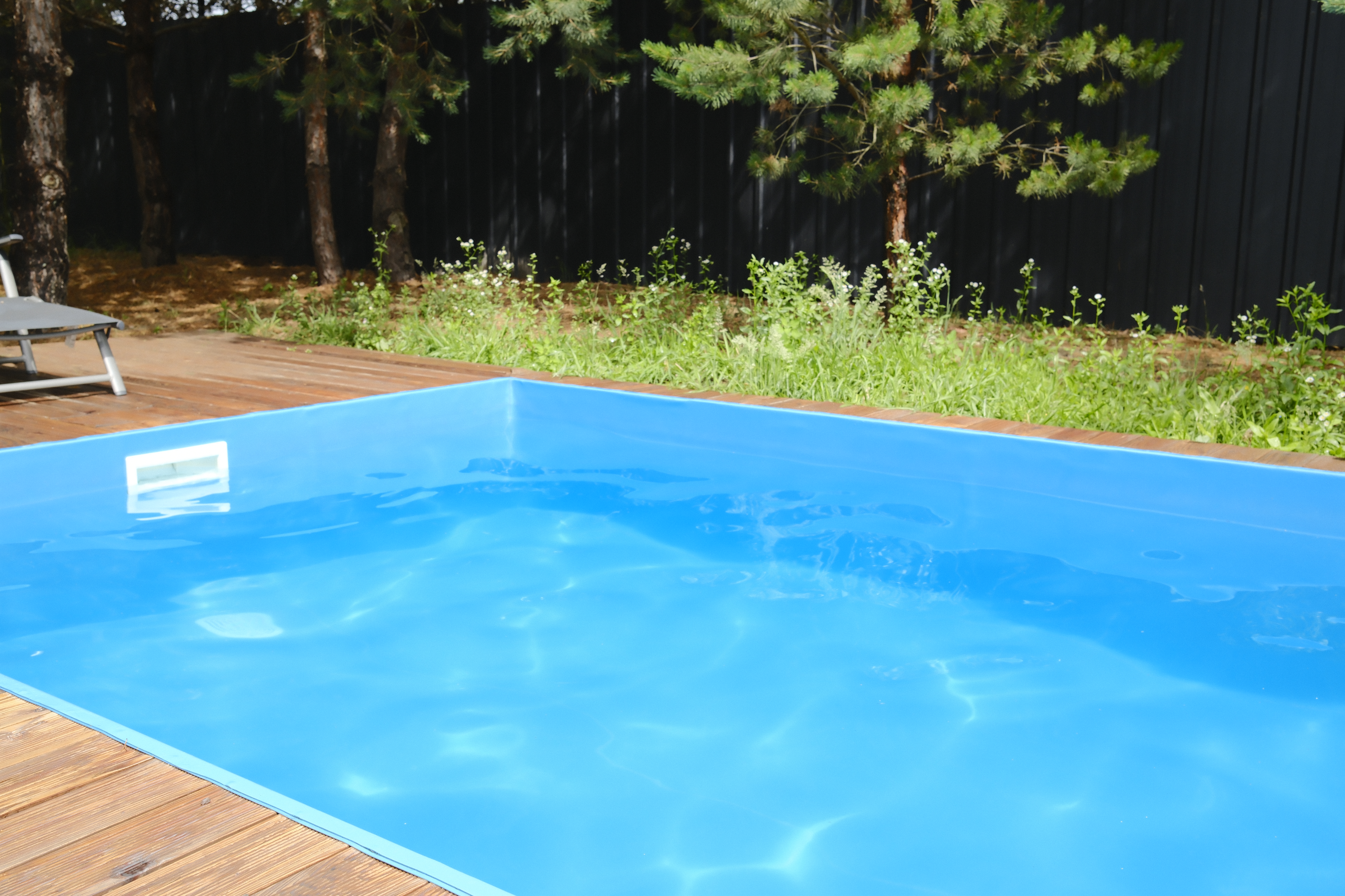 Clear blue water fills a backyard swimming pool surrounded by a wooden deck and green trees on a sunny summer day while a lounge chair sits nearby on the grass
